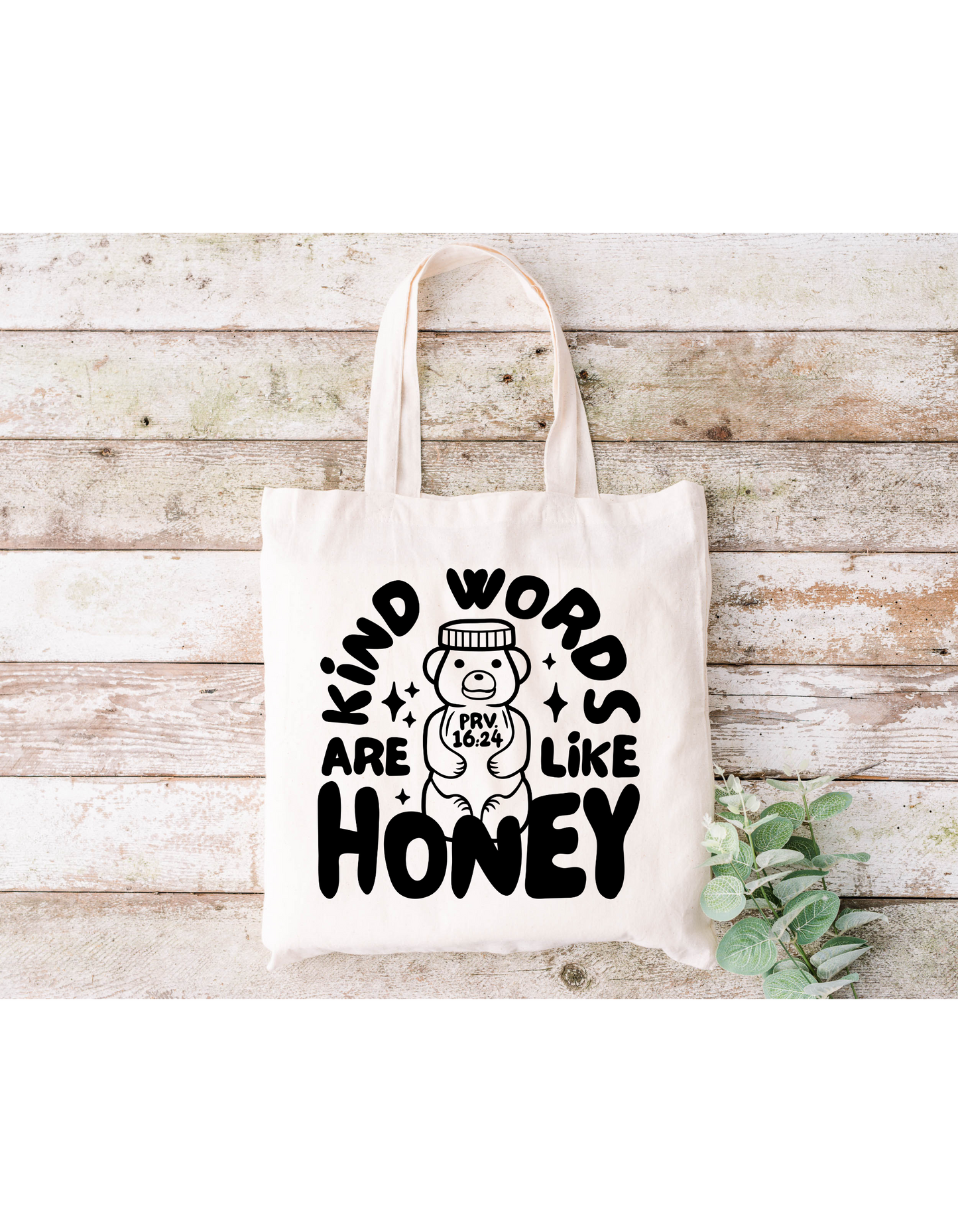 The Kind Words Are Like Honey Canvas Bag is a beige cotton tote featuring the text Kind words are like honey in black on a wooden background. A bear drawing with Prov 16:24 on its belly and green leaves accent it, ideal for bible study or casual outings.