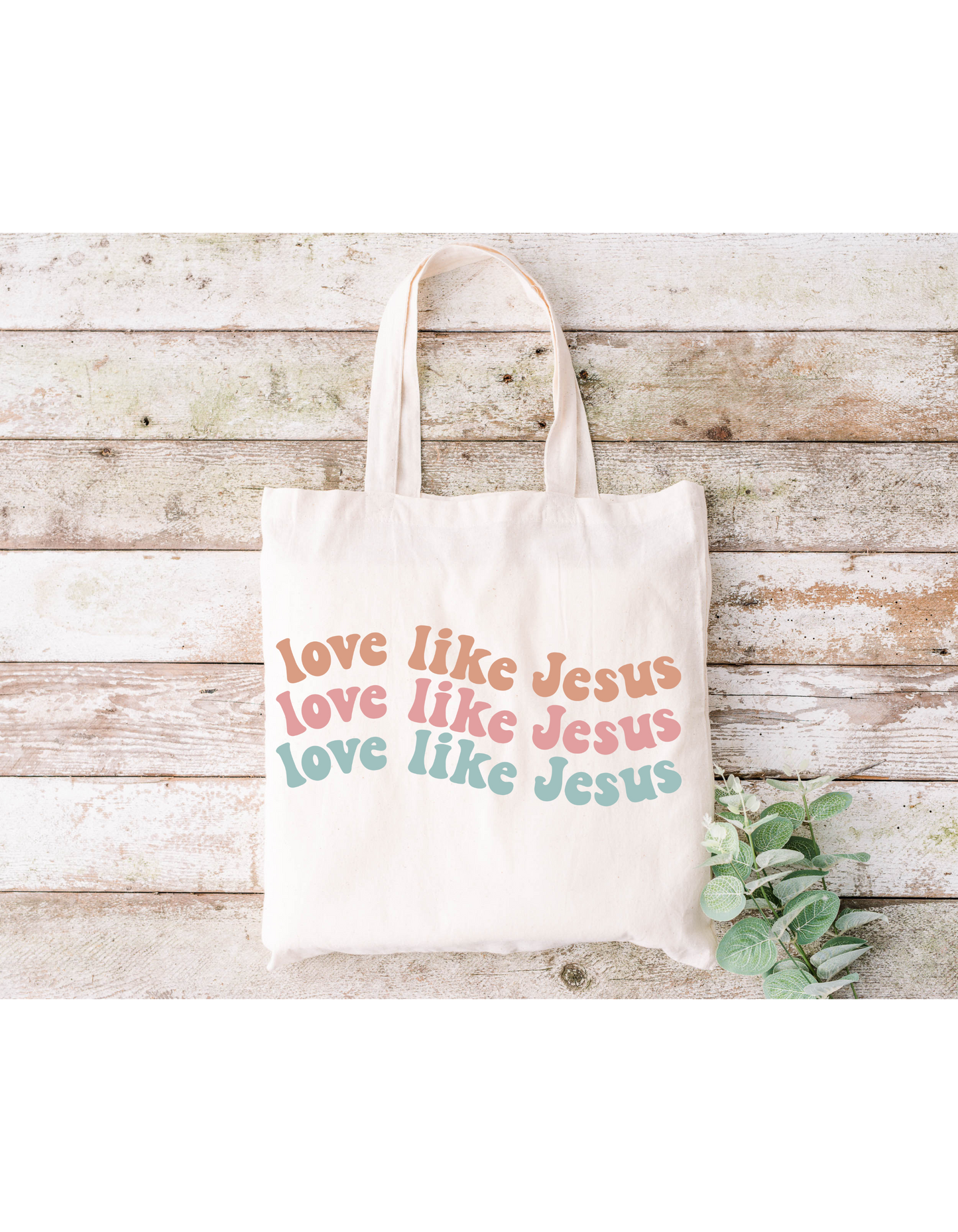 The Love Like Jesus Canvas Bag is a cream cotton tote featuring love like Jesus repeated in pink, red, and teal. Ideal for bible study, it sits on a rustic wooden surface adorned with greenery.