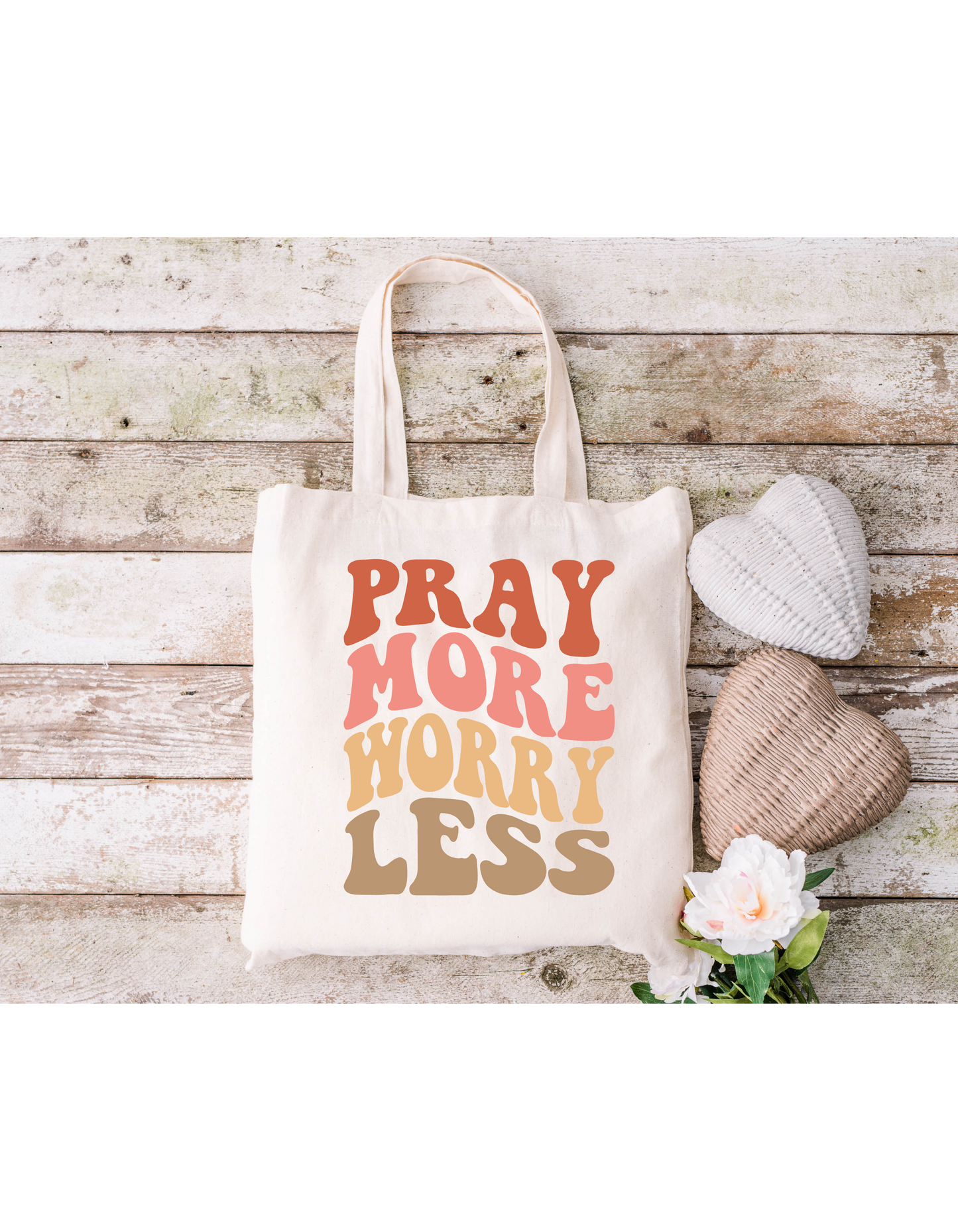 A Pray More Worry Less Canvas Bag sits on a rustic wooden surface alongside two heart-shaped cushions and a white flower, perfect for bible study essentials.
