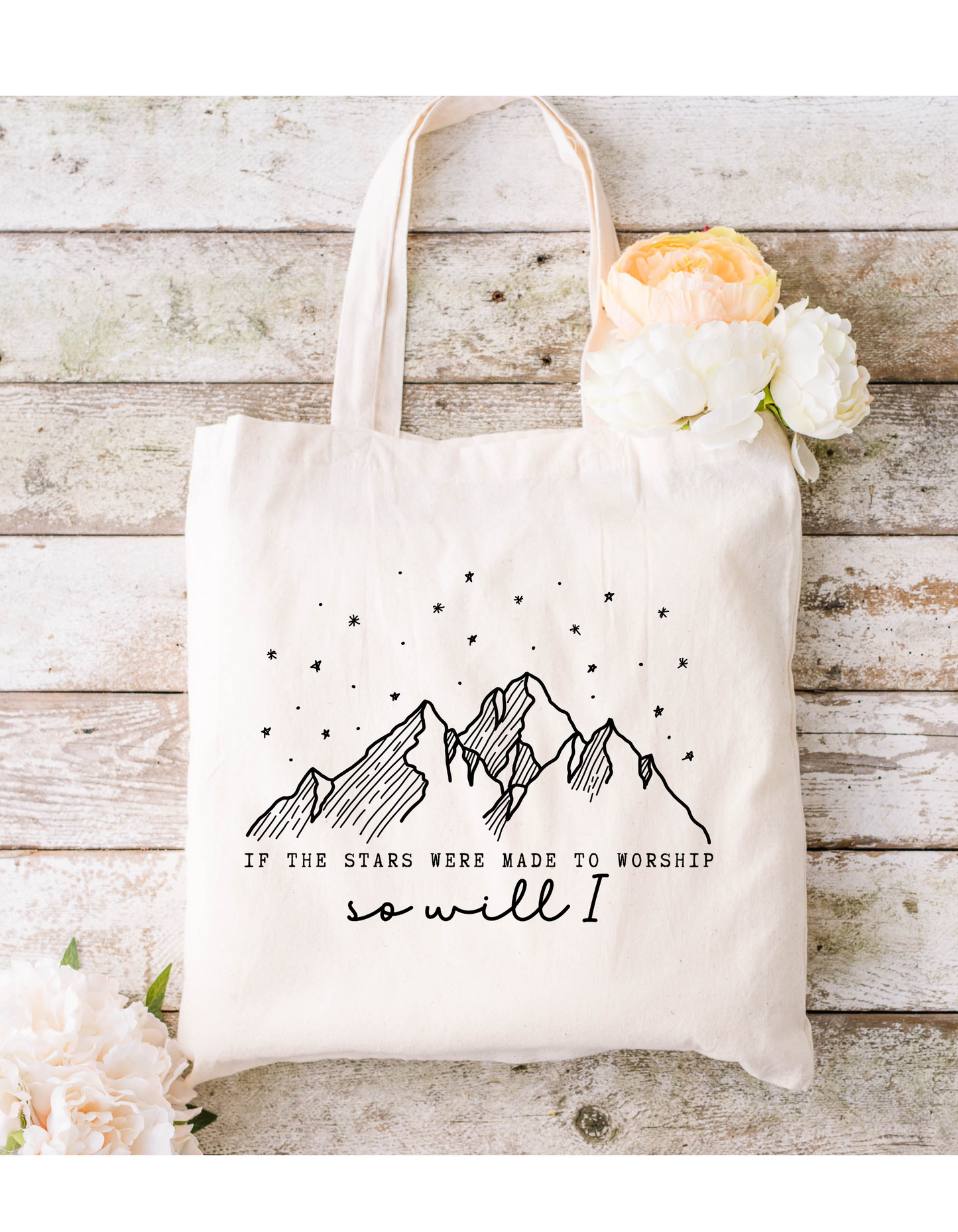 Featuring a rustic wooden background, the If the Stars Were Made to Worship Canvas Bag has a black line mountain and star drawing with text that reads: If the stars were made to worship, so will I. Adorned with white and peach flowers, its ideal for bible study or daily use.