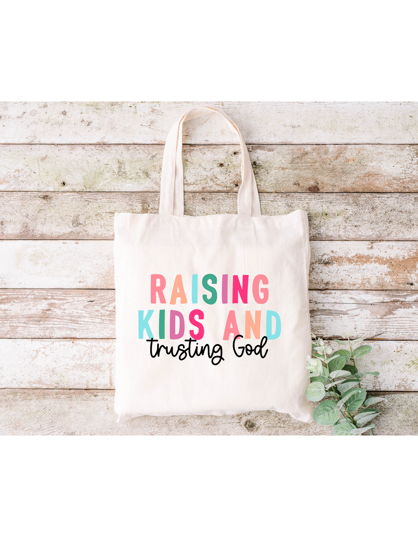 The Raising Kids and Trusting God (Colorful) Canvas Bag, a beige cotton tote with nylon webbed straps, displays its message in vibrant lettering. Stylishly placed on wood and accented by green leaves, it’s perfect for your next bible study session.