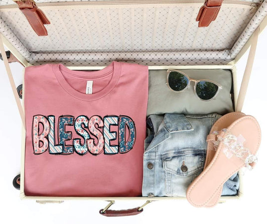 An open suitcase contains a pink buttery soft Blessed Short Sleeve Tee with BLESSED in floral patterns, sunglasses, a light denim jacket, olive green pants, and beige sandals with clear straps.