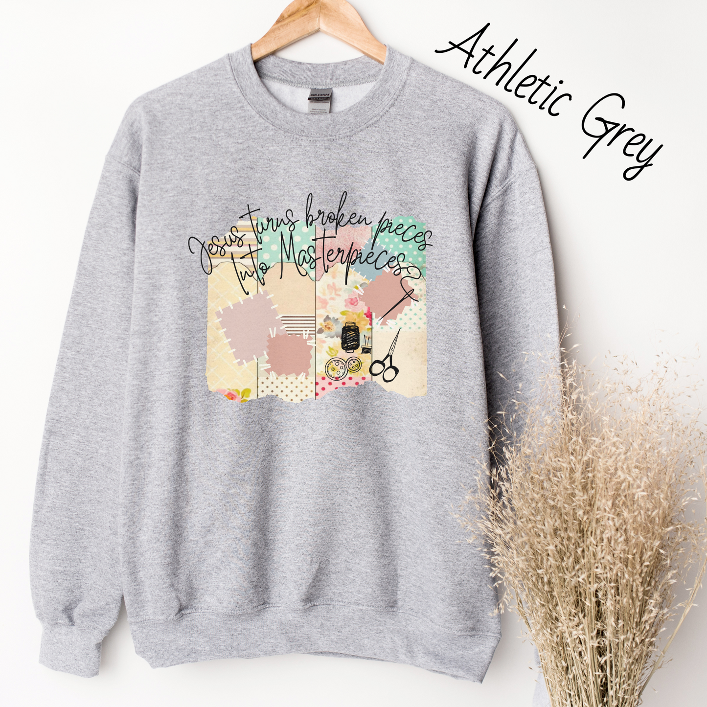 Light grey sweatshirt with multicolored quilt design with the words "Jesus turns broken pieces into masterpieces written across the top in black cursive script.