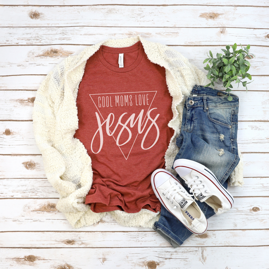 The Cool Moms Love Jesus Short Sleeve Tee in red, crafted from buttery soft Airlume combed cotton, is paired with a white cardigan, distressed jeans, and white sneakers. Nearby is a small green plant. All items are displayed on a light wooden surface.
