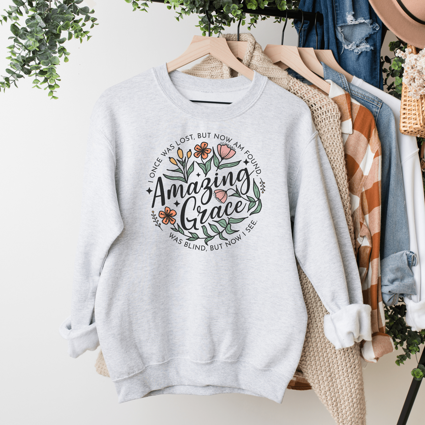 Words "Amazing Grace" with florals around and "I once was lost but now am found, was blind but now I see" in a circle around the main design. Design is on an ash colored sweatshit hanging on a rack.