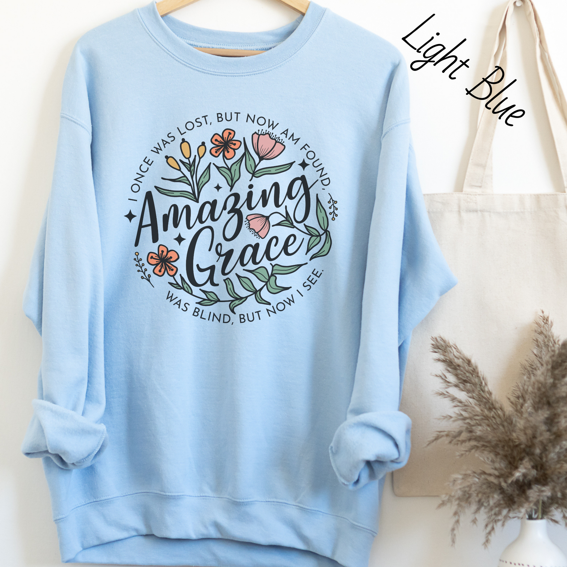 Words "Amazing Grace" with florals around and "I once was lost but now am found, was blind but now I see" in a circle around the main design. Design is on a light blue colored sweatshit.