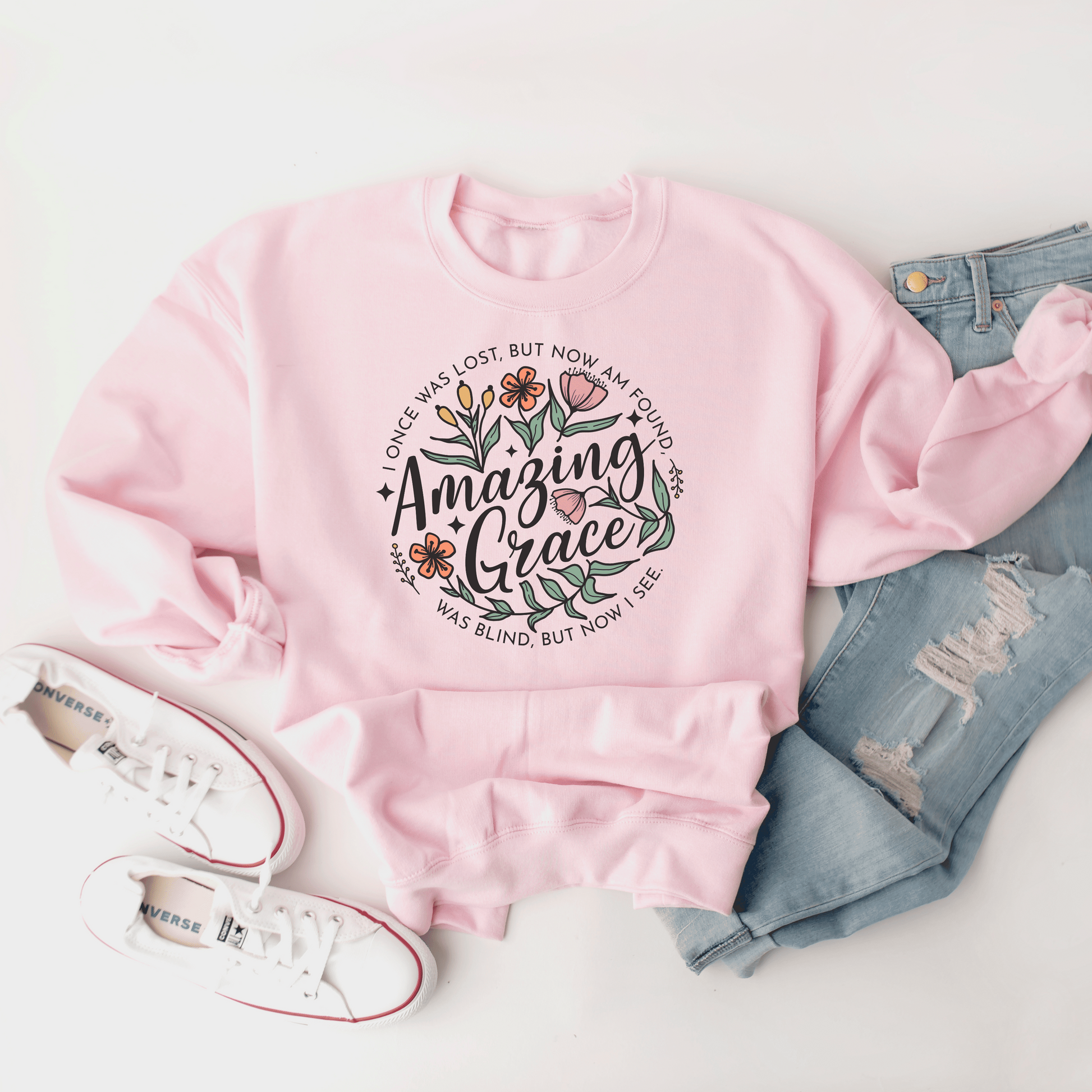 Words "Amazing Grace" with florals around and "I once was lost but now am found, was blind but now I see" in a circle around the main design. Design is on a light pink colored sweatshit.