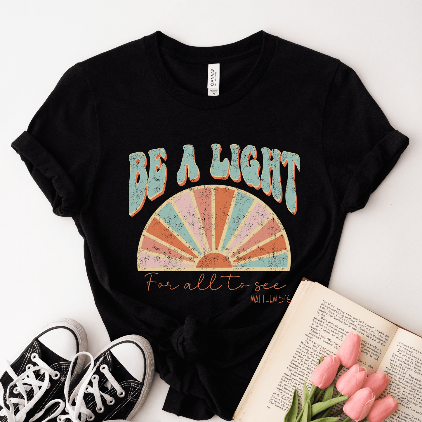 A buttery soft black T-shirt, the Be A Light Short Sleeve Tee, made with Airlume combed cotton, features Be a Light in distressed blue and peach letters alongside a colorful semi-circle. Below it reads For all to see, Matthew 5:16. Nearby are black sneakers, pink tulips, and an open book.