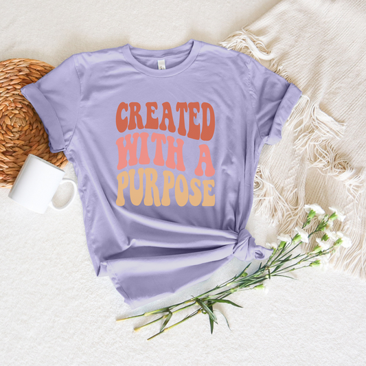 The Created With A Purpose Retro Style Short Sleeve Tee in lavender, made from buttery soft Airlume cotton and styled with a bottom knot, is paired with a white mug, a woven placemat, beige fringed cloth, and white flowers on a light background.
