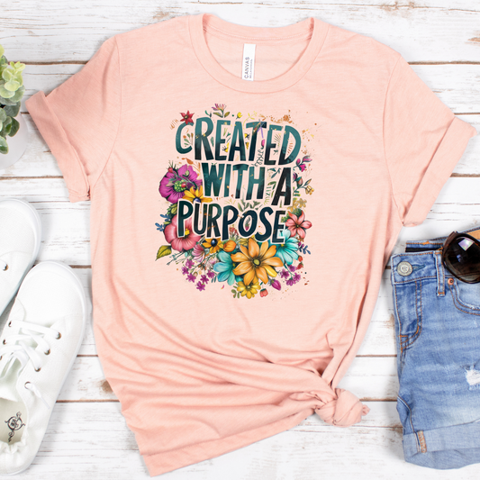 The Created With A Purpose Floral Style Short Sleeve Tee is peach-colored with multicolored flowers on buttery soft Airlume combed cotton, resting against a white rustic wood backdrop with sneakers, denim, and sunglasses.