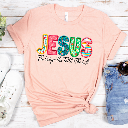 The Jesus The Way The Truth The Life (Spring Floral) Short Sleeve Tee in peach, crafted from soft Airlume cotton, showcases JESUS in colorful letters above The Way + The Truth + The Life. Styled flat with denim shorts, white sneakers, and sunglasses on a white wood surface.