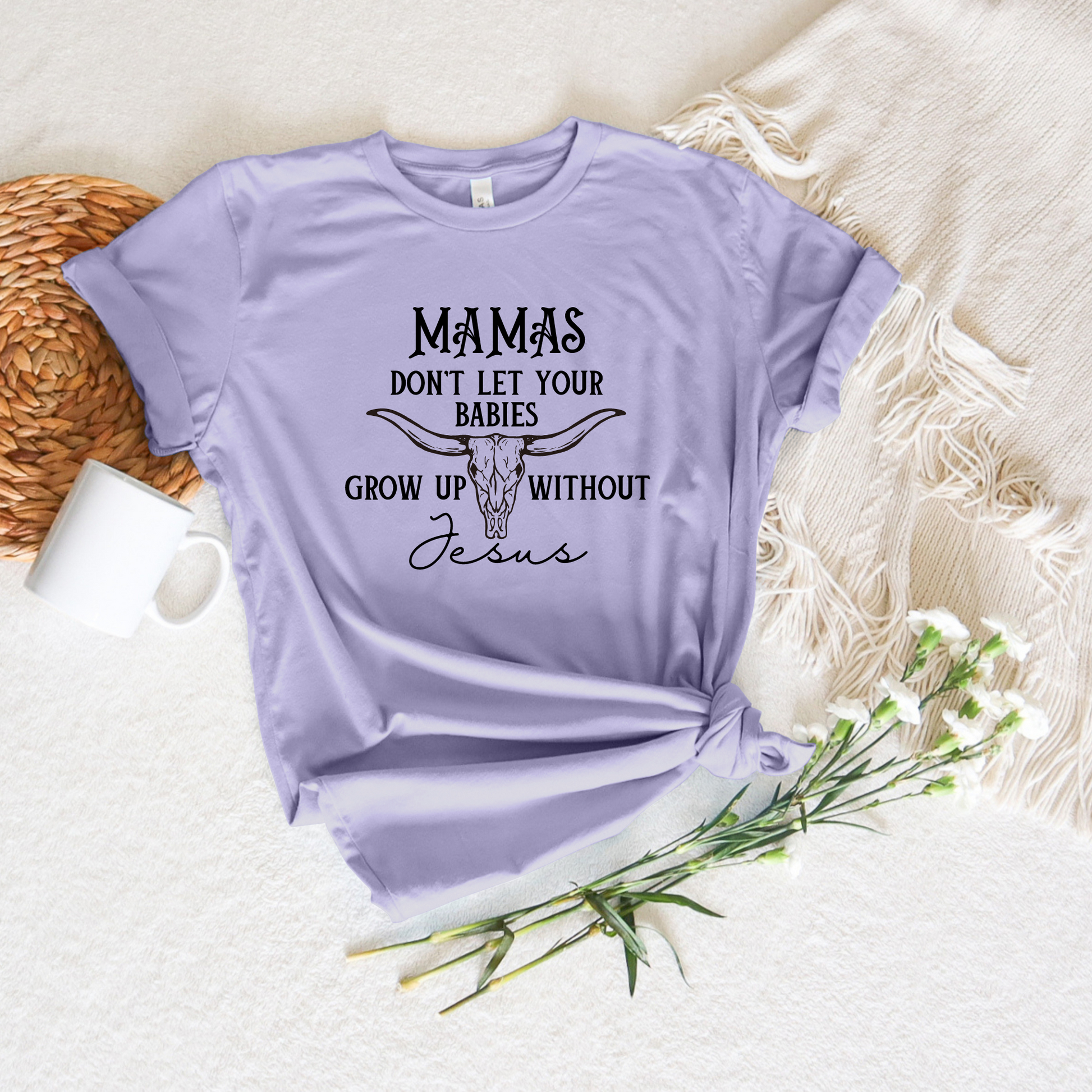 Lilac Mamas Dont Let Your Babies Grow Up Without Jesus short sleeve tee with a bull skull design on a textured backdrop, paired with a white mug, woven basket, and delicate white flowers.
