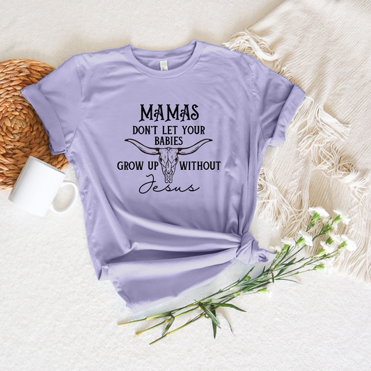 Lilac Mamas Dont Let Your Babies Grow Up Without Jesus short sleeve tee with a bull skull design on a textured backdrop, paired with a white mug, woven basket, and delicate white flowers.