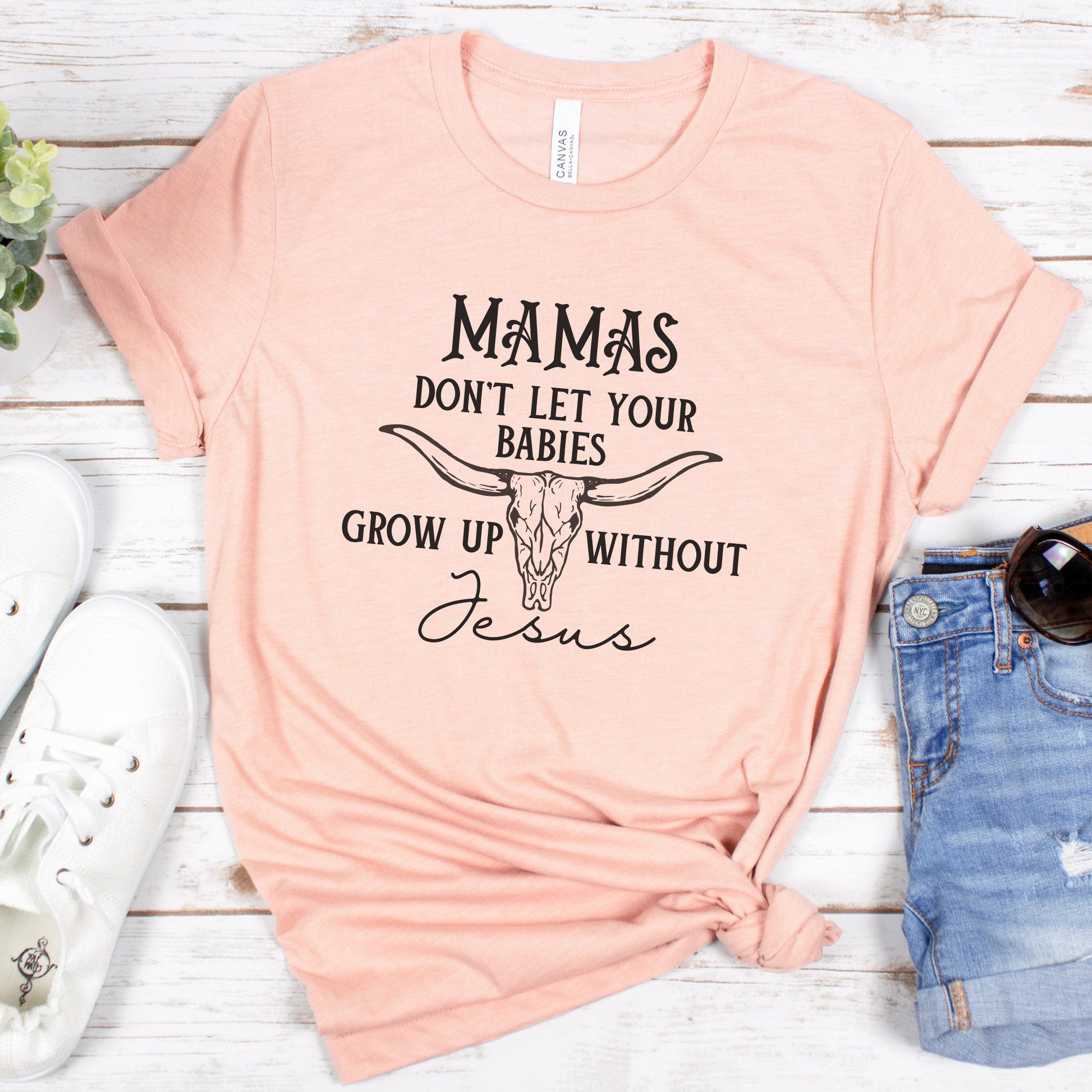 The Mamas Dont Let Your Babies Grow Up Without Jesus Short Sleeve Tee, in a peach shade with Mamas dont let your babies grow up without Jesus text and a longhorn graphic, pairs perfectly with white sneakers and denim shorts. Styled on wood with a small potted plant, it exudes warmth and style.