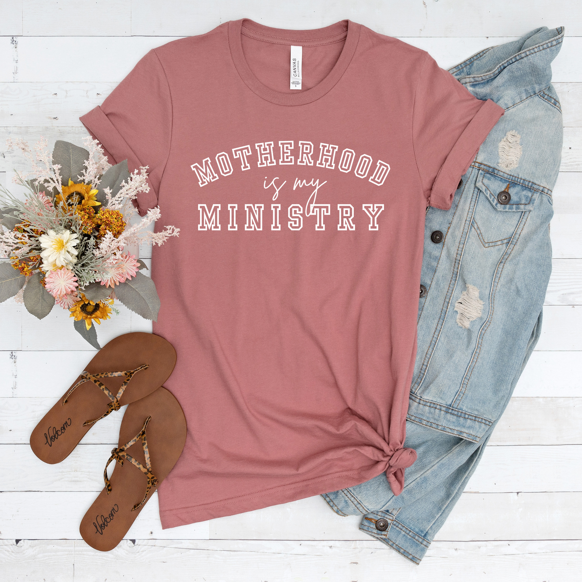 A buttery soft Motherhood Is My Ministry Short Sleeve Tee in pink rests on a white wooden floor. Brown sandals, a denim jacket, and a bouquet of flowers beside it add grace to your daily walk with God.