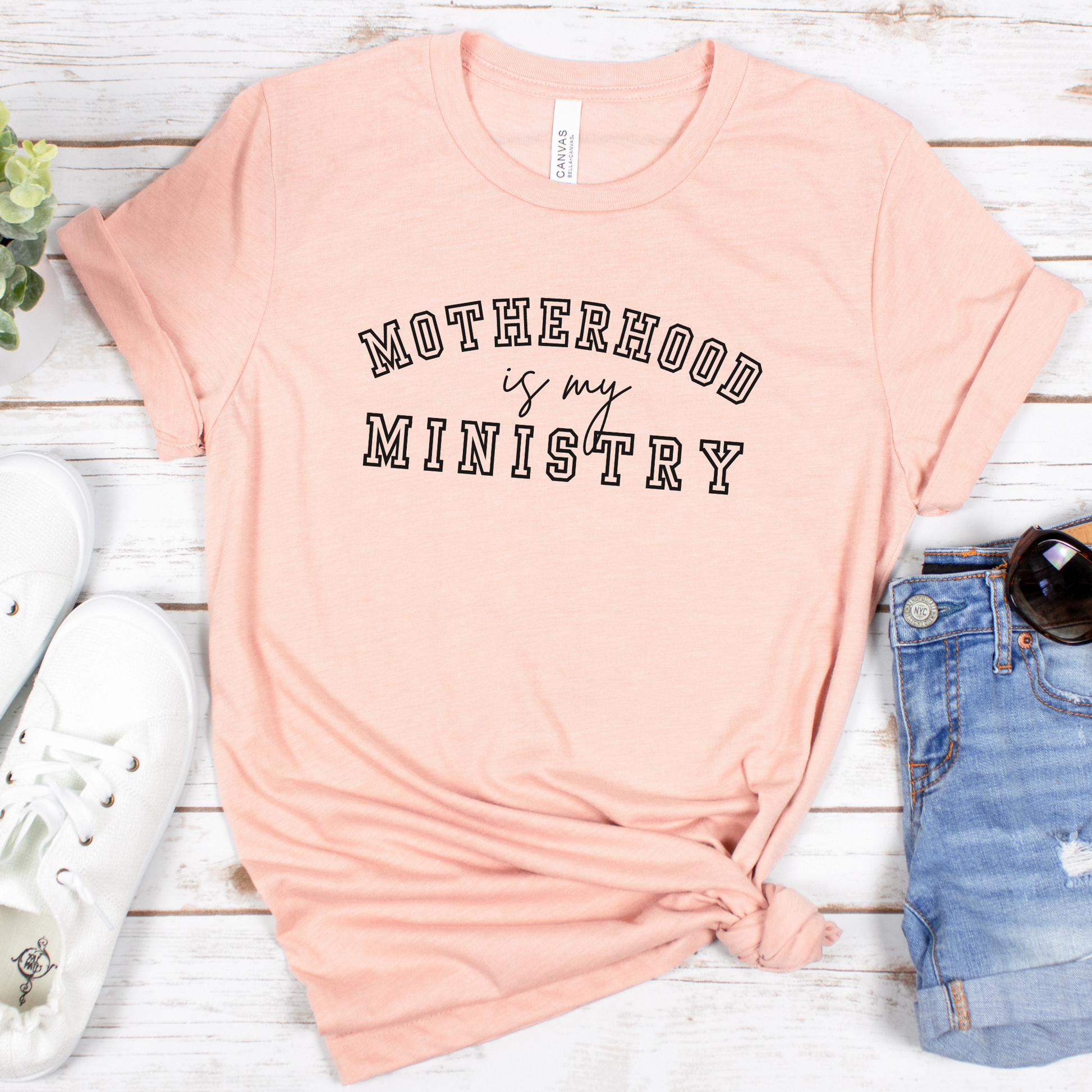 The Motherhood Is My Ministry Short Sleeve Tee in delicate pink, with bold black lettering, effortlessly pairs with white sneakers, denim shorts, chic sunglasses, and is styled next to a vibrant small plant on a white wooden surfaceâ€”perfect for showcasing casual grace and devotion.
