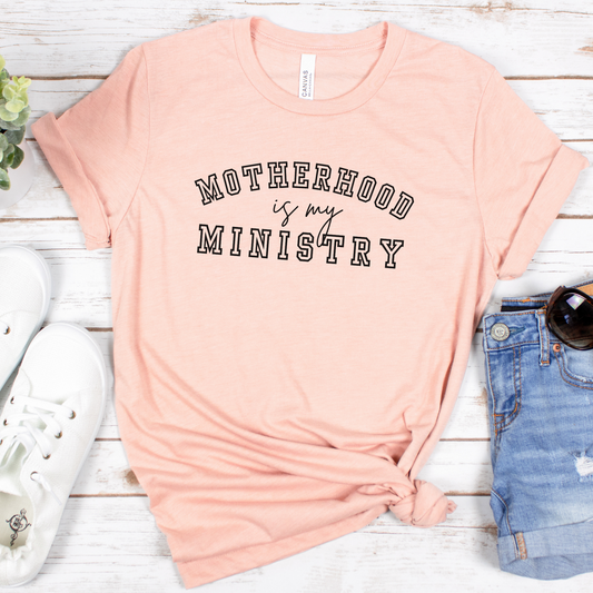 The Motherhood Is My Ministry Short Sleeve Tee in delicate pink, with bold black lettering, effortlessly pairs with white sneakers, denim shorts, chic sunglasses, and is styled next to a vibrant small plant on a white wooden surfaceâ€”perfect for showcasing casual grace and devotion.