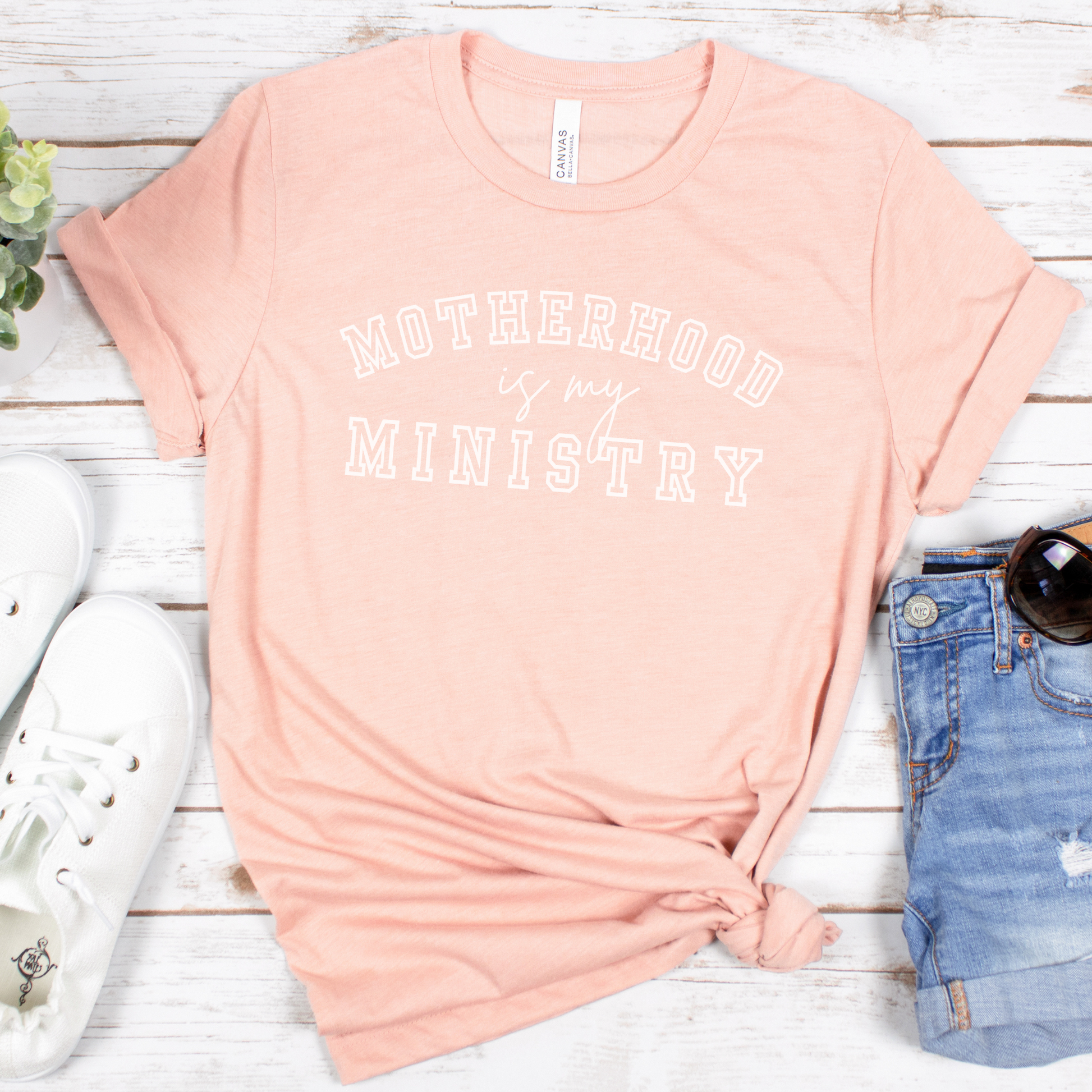 A buttery soft, peach-colored Motherhood Is My Ministry short sleeve tee lies gracefully on a white wooden surface, paired with white sneakers, denim shorts, sunglasses, and a small plant. The knotted tee exudes both comfort and devotion.