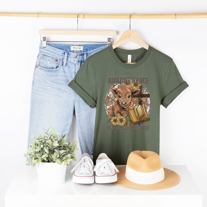 A circle with a highland calf, sunflowers, a cross, and an open bible with the words "Amazing Grace" above and "How Sweet The Sound" below. Design is on a pine colored shirt.