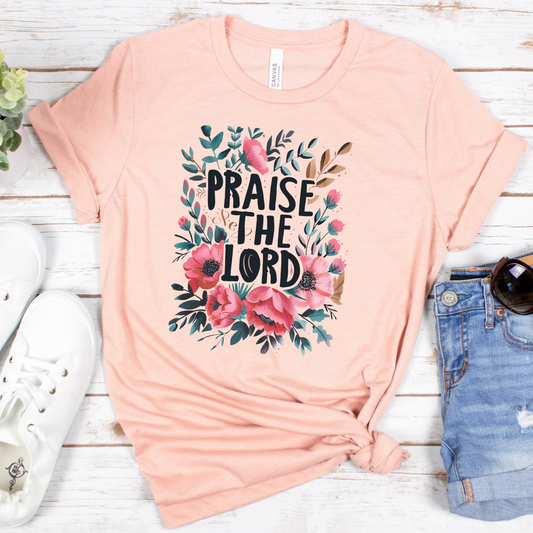 The Praise The Lord Short Sleeve Tee in peach, made from soft Airlume cotton, boasts a floral design of pink and red flowers with green leaves. Styled on a white wooden surface, its ideally matched with white sneakers, denim shorts, and sunglasses for a breezy look.