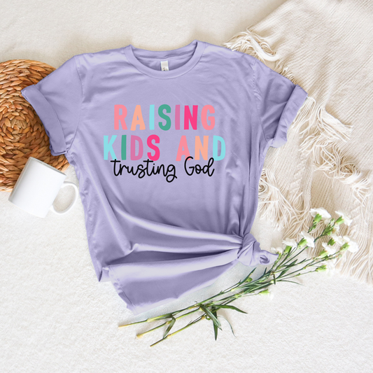 The Raising Kids and Trusting God Short Sleeve Tee in lavender, crafted from soft Airlume cotton, features the colorful phrase against a cream woven blanket, accompanied by a white mug, wicker mat, and delicate white flowers.