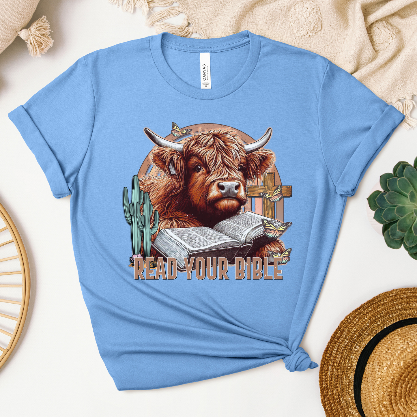 The Read Your Bible Highland Cow Short Sleeve Tee in light blue features a highland cow reading a Bible amidst a cactus, cross, and butterflies, with the text Read Your Bible. Made from soft Airlume cotton, its styled with a knot.