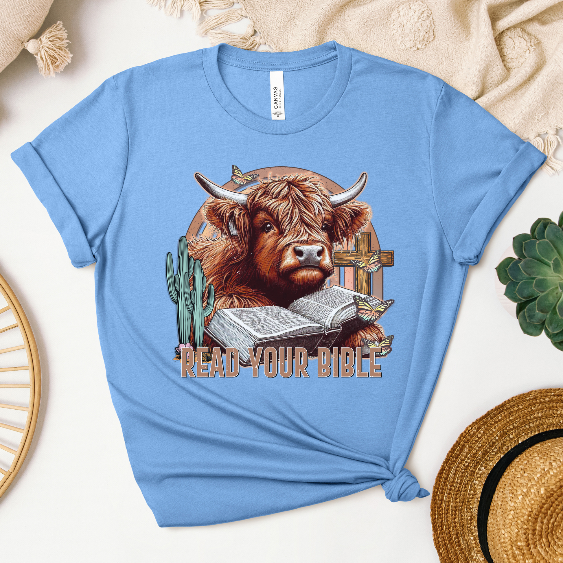 The Read Your Bible Highland Cow Short Sleeve Tee in light blue features a highland cow reading a Bible amidst a cactus, cross, and butterflies, with the text Read Your Bible. Made from soft Airlume cotton, its styled with a knot.