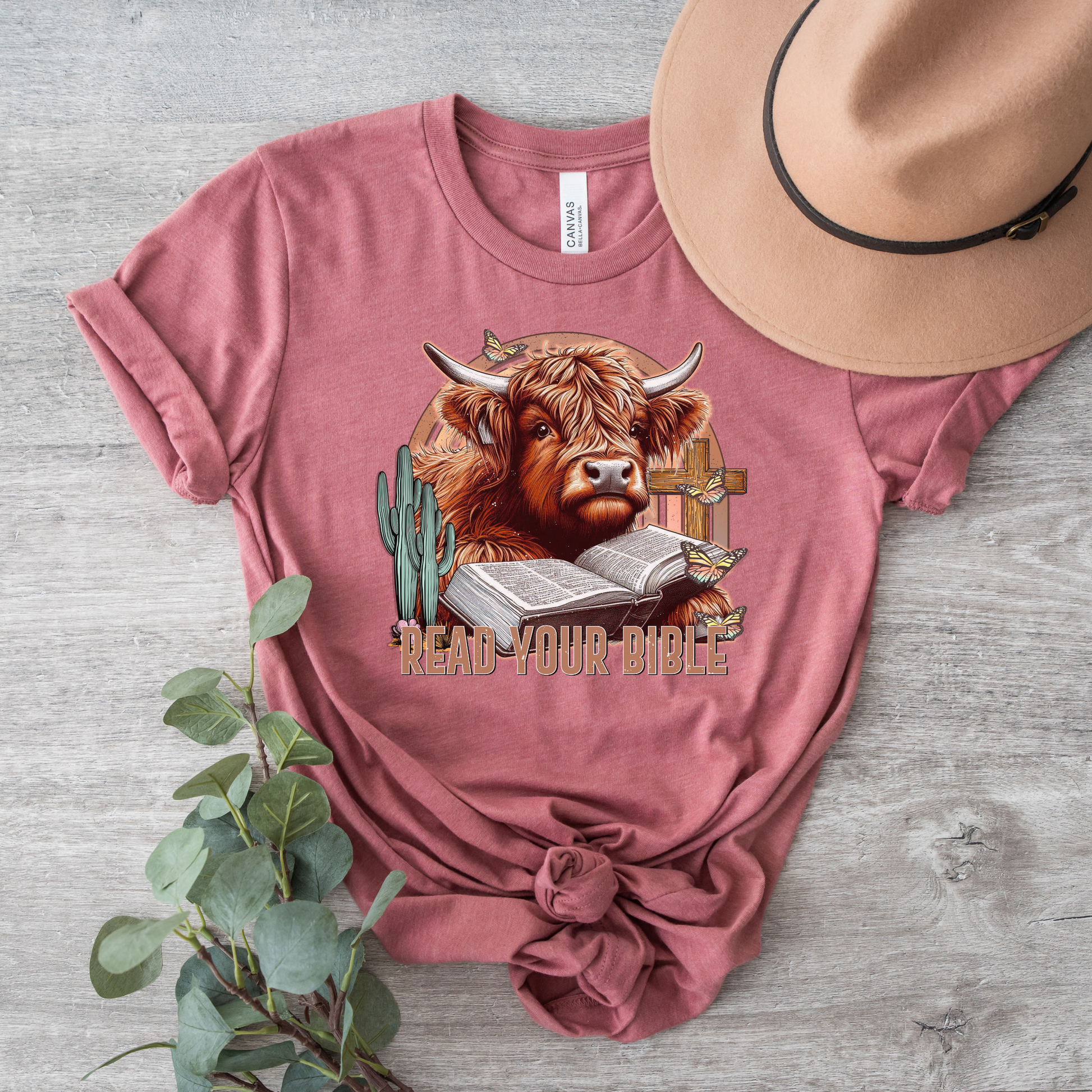 The Read Your Bible Highland Cow Short Sleeve Tee is a pink T-shirt made from buttery soft Airlume cotton, displaying a highland cow, cactus, open Bible, and cowboy hat. Its enhanced by the text Read Your Bible, with a tan hat and greenery accenting its divine display on wood.