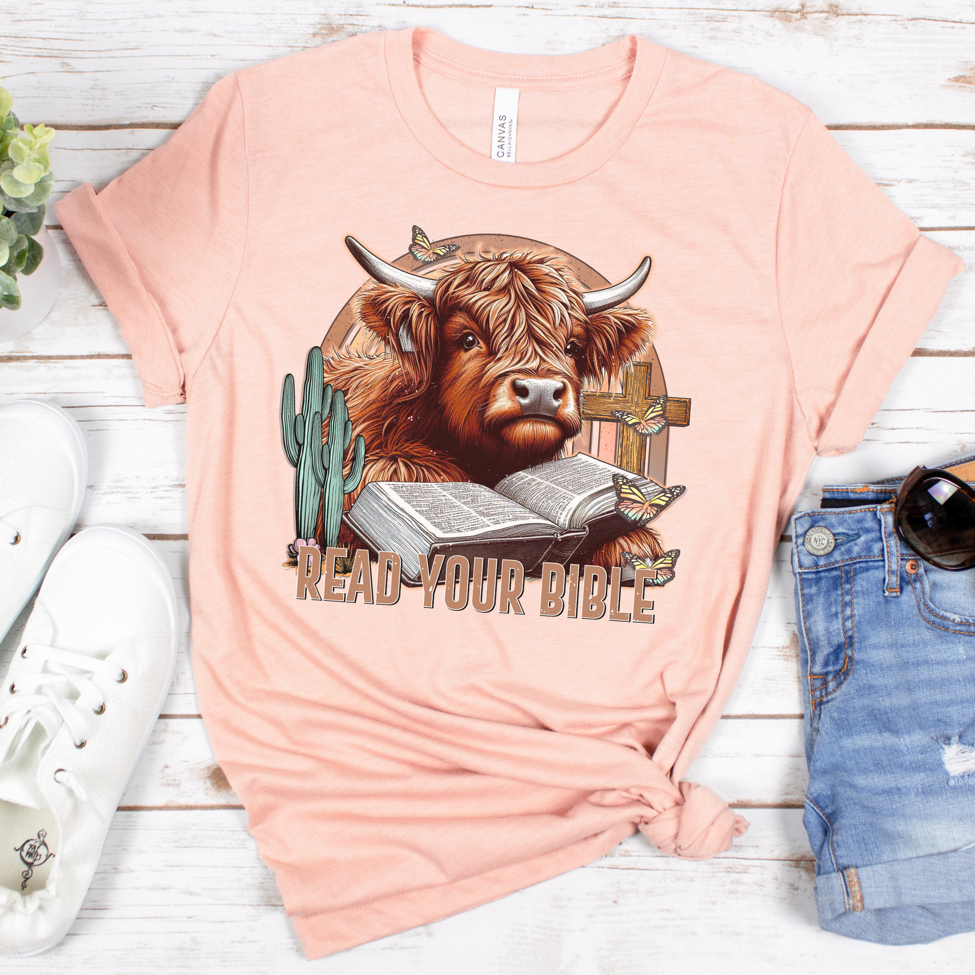 The Read Your Bible Highland Cow Short Sleeve Tee is a pink Airlume cotton T-shirt with a cow wearing glasses reading the Bible amid butterflies and a cactus. READ YOUR BIBLE is displayed above the design. It sits on a wooden surface next to denim shorts and white sneakers.
