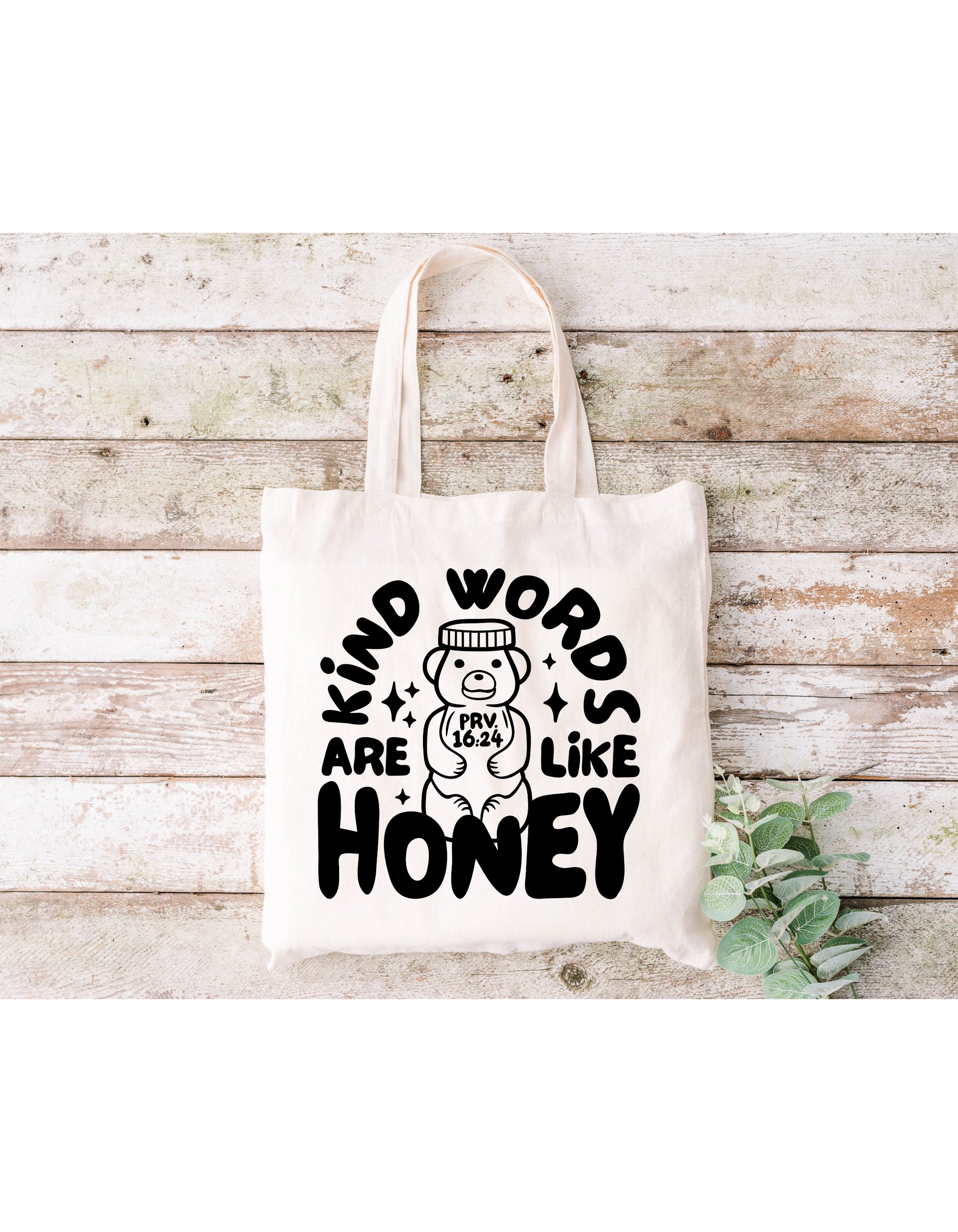 The Kind Words Are Like Honey Canvas Bag is a beige cotton tote featuring the text Kind words are like honey in black on a wooden background. A bear drawing with Prov 16:24 on its belly and green leaves accent it, ideal for bible study or casual outings.