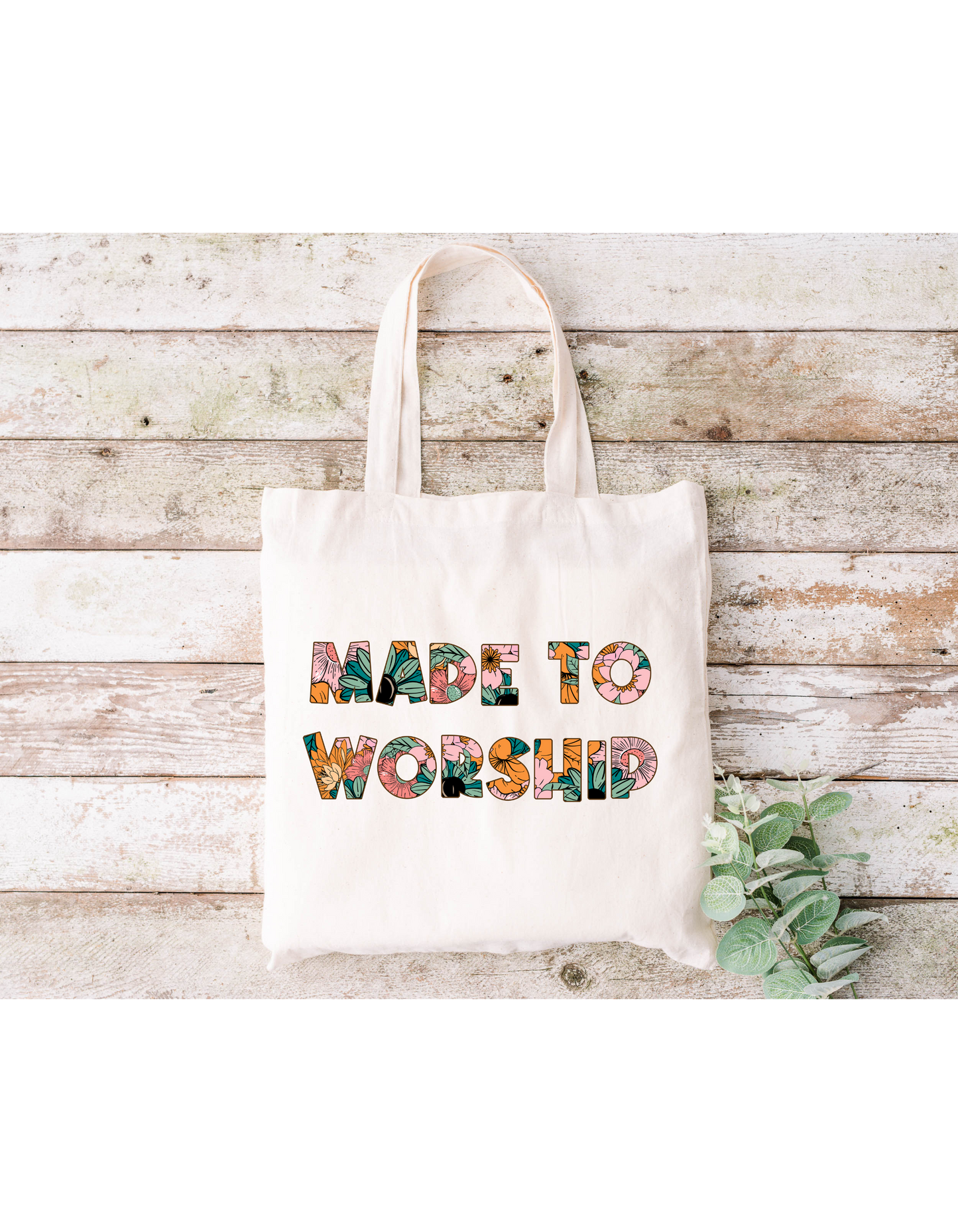 A Made to Worship (Floral) Canvas Bag rests on a rustic wooden surface with a small leafy branch, adding a touch of natureâ€”ideal for bible study or everyday essentials.