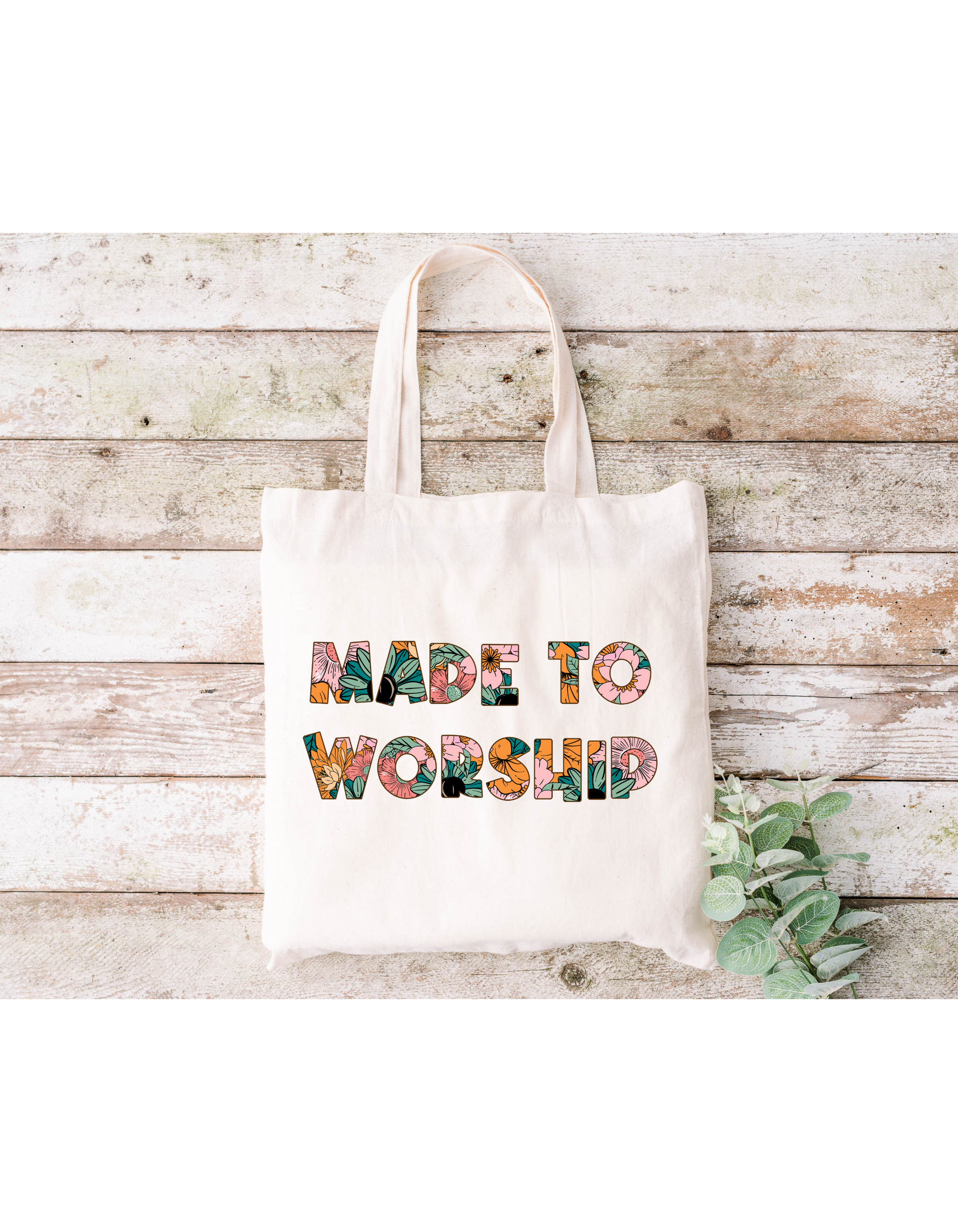 A Made to Worship (Floral) Canvas Bag rests on a rustic wooden surface with a small leafy branch, adding a touch of natureâ€”ideal for bible study or everyday essentials.