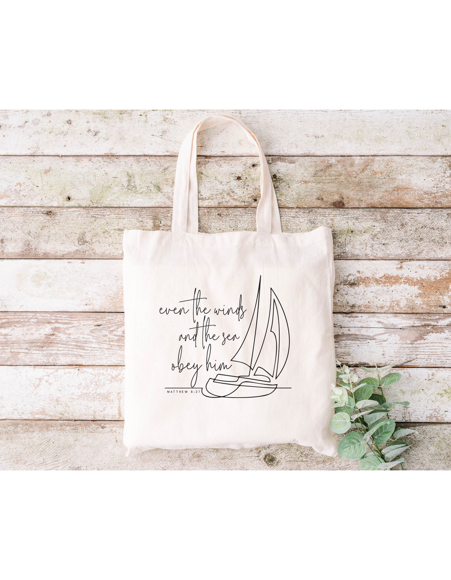 The Even the Winds and Seas Obey Him canvas bag, made of beige cotton, showcases a sailboat outline with the phrase. Its ideal for Bible study sessions and is displayed on wooden planks with green leaves in the lower-right corner.