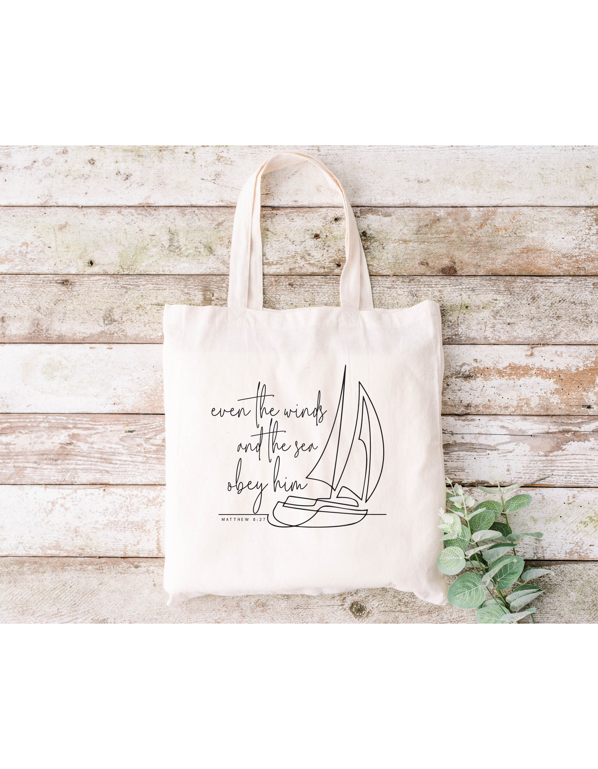 The Even the Winds and Seas Obey Him canvas bag, made of beige cotton, showcases a sailboat outline with the phrase. Its ideal for Bible study sessions and is displayed on wooden planks with green leaves in the lower-right corner.