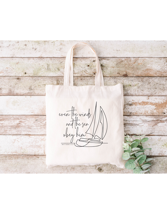 The Even the Winds and Seas Obey Him canvas bag, made of beige cotton, showcases a sailboat outline with the phrase. Its ideal for Bible study sessions and is displayed on wooden planks with green leaves in the lower-right corner.