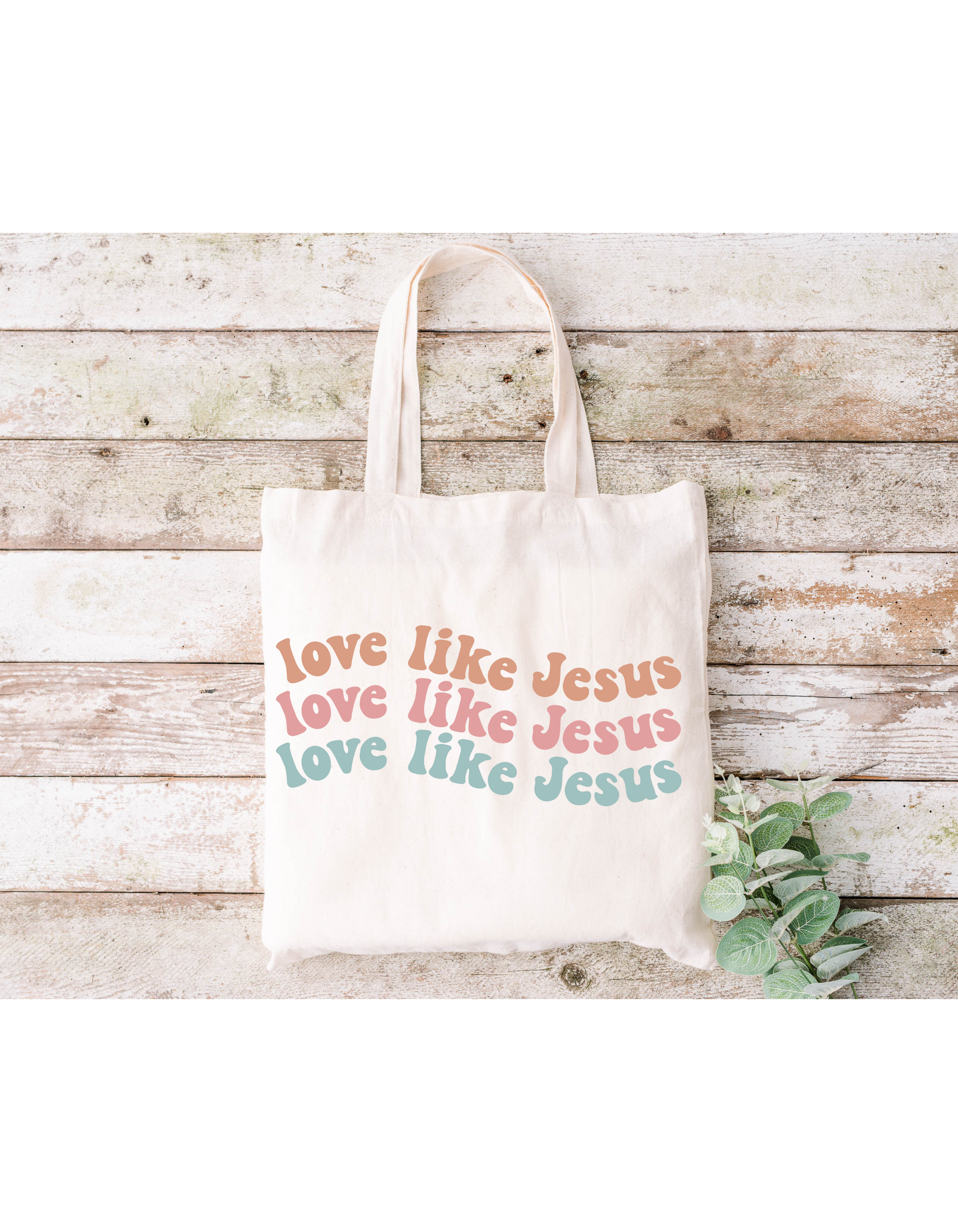 The Love Like Jesus Canvas Bag is a cream cotton tote featuring love like Jesus repeated in pink, red, and teal. Ideal for bible study, it sits on a rustic wooden surface adorned with greenery.
