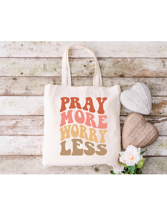 A Pray More Worry Less Canvas Bag sits on a rustic wooden surface alongside two heart-shaped cushions and a white flower, perfect for bible study essentials.