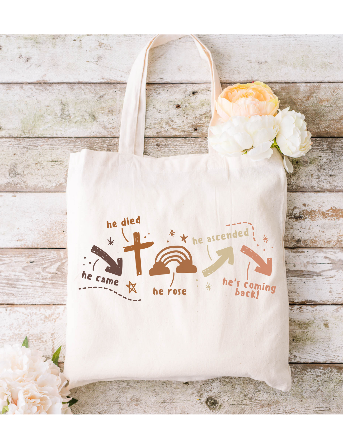 The Hes Coming Back Canvas Bag is a beige cotton tote featuring Christian designs of a cross, tomb, and arrows with the text: he died, he came, he rose, he ascended, hes coming back! Perfect for Bible study sessions and displayed on a wooden surface with white and peach flowers.