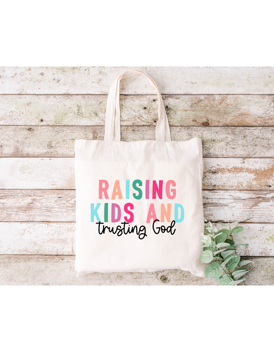 The Raising Kids and Trusting God (Colorful) Canvas Bag, a beige cotton tote with nylon webbed straps, displays its message in vibrant lettering. Stylishly placed on wood and accented by green leaves, itâ€™s perfect for your next bible study session.
