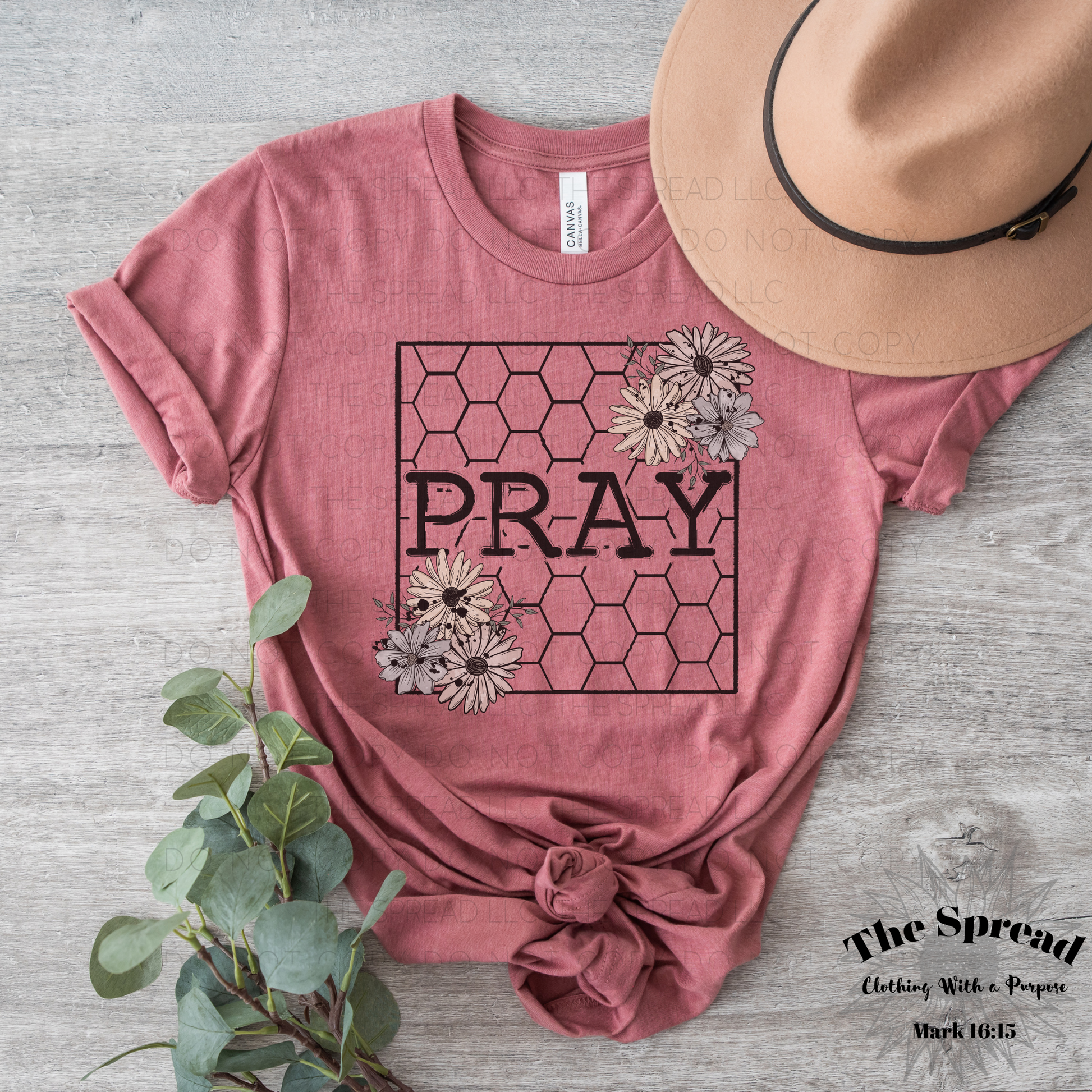 The Pray Honeycomb Short Sleeve Tee is a mauve t-shirt made from Airlume combed cotton featuring PRAY inside a hexagon with flowers. Styled with a twisted knot, it lies beside a beige hat and greenery on wood, providing comfort like buttery soft tees.