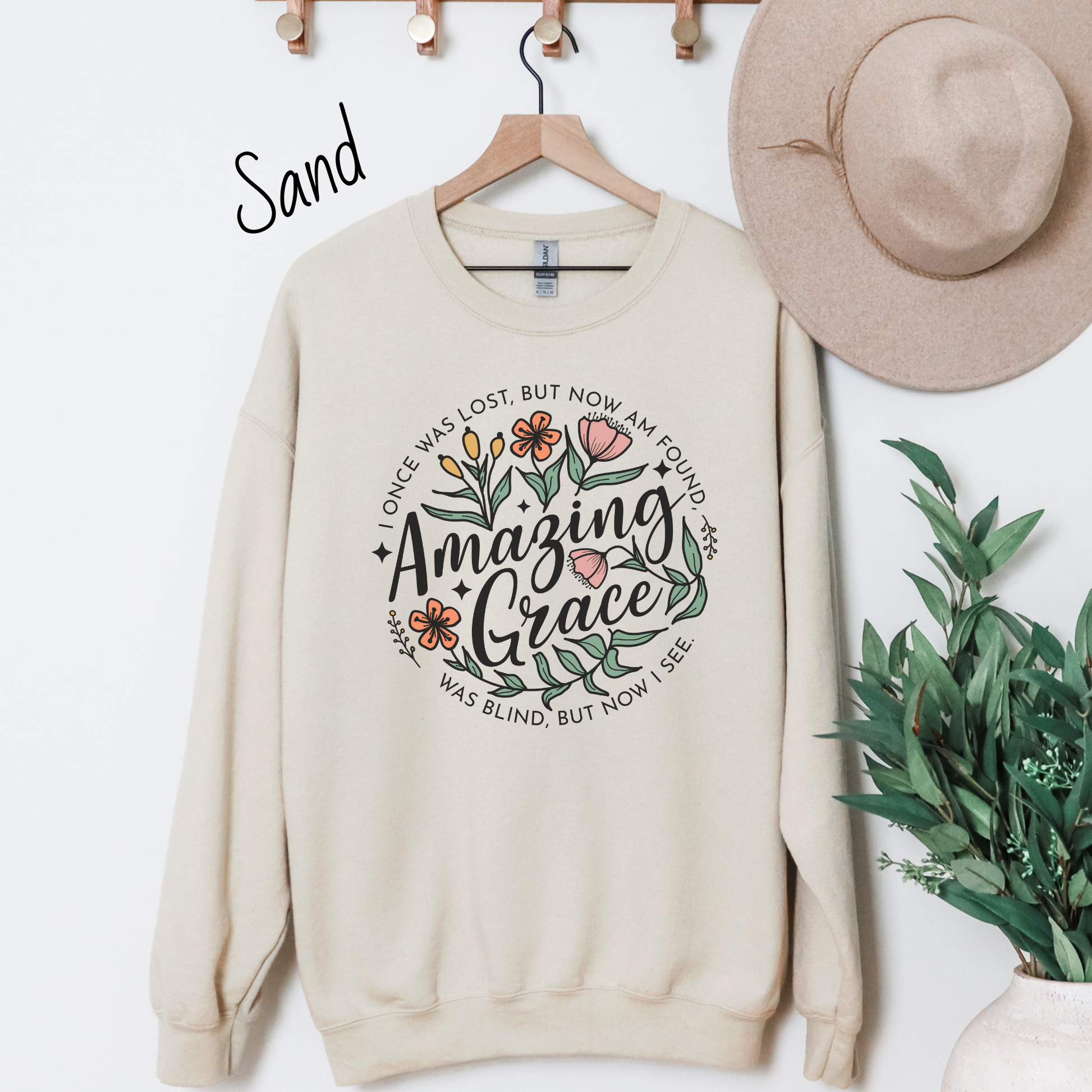 Words "Amazing Grace" with florals around and "I once was lost but now am found, was blind but now I see" in a circle around the main design. Design is on a sand colored sweatshit hanging on a rack.